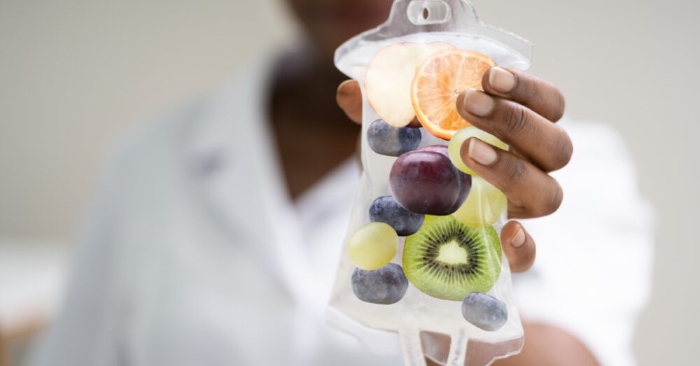 Close-up of a hand holding an IV bag filled with colorful fruits including kiwi, berries, and citrus slices against a soft blurred background