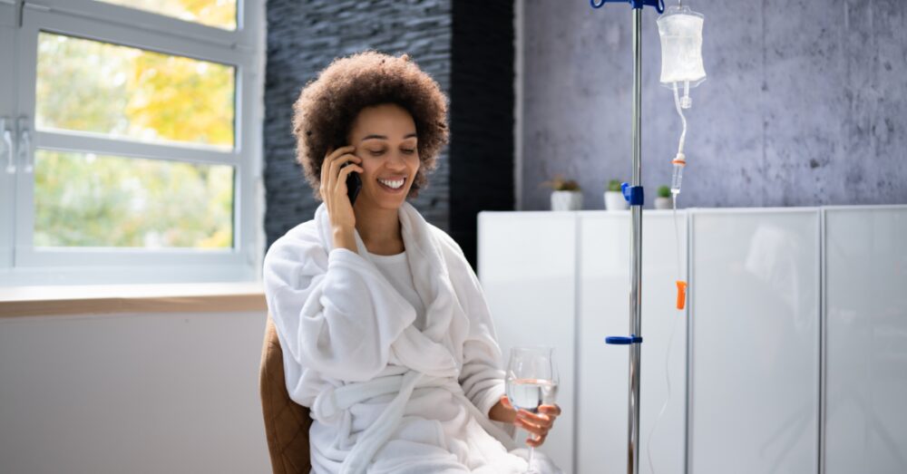 Woman in a white robe smiling and talking on the phone while receiving an IV drip treatment in a bright modern room